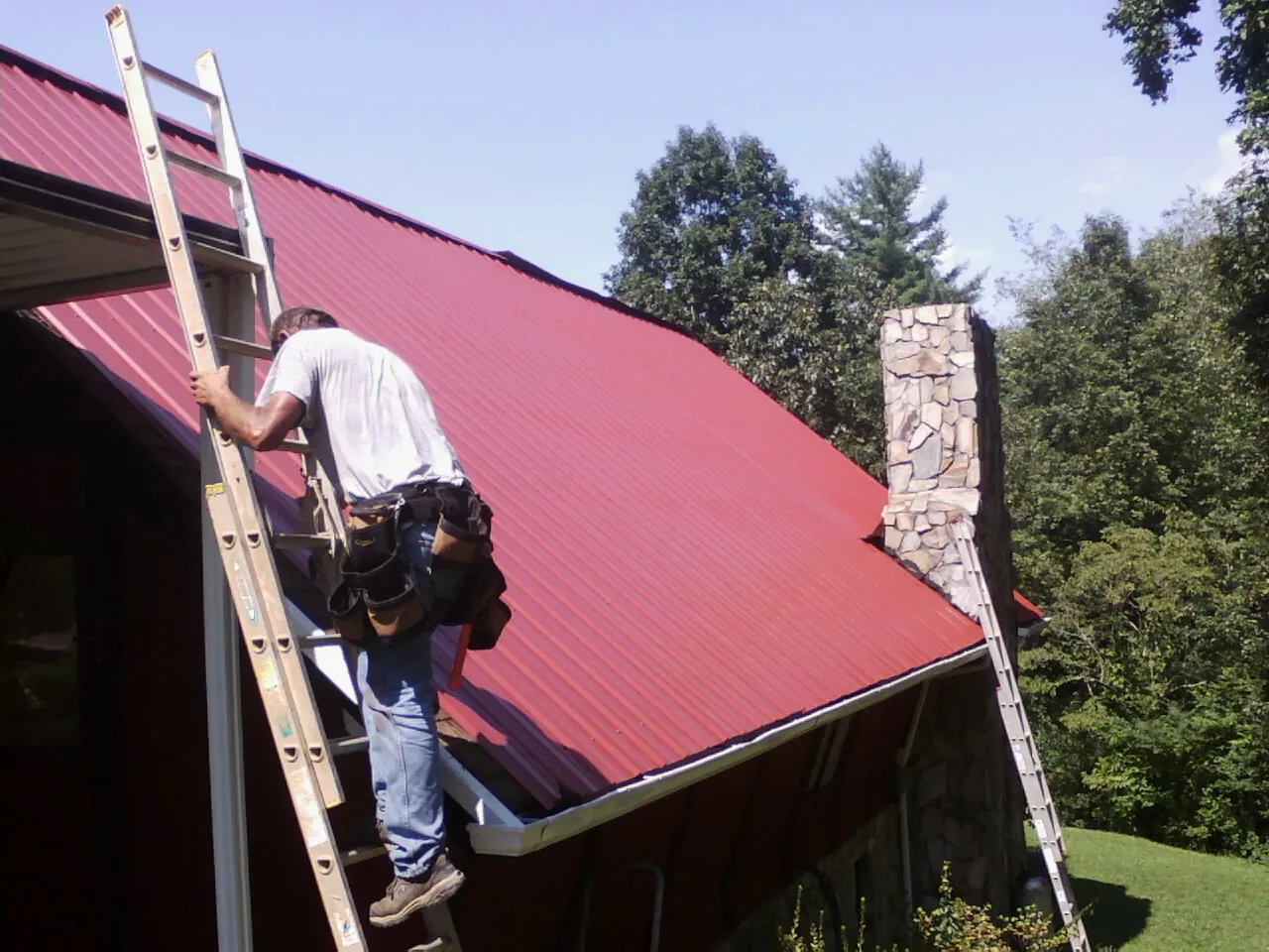 Roofer on a ladder beside a red metal roof and stone chimney — Soffit & Fascia Repair by Roofing Brothers in Saint Inigoes